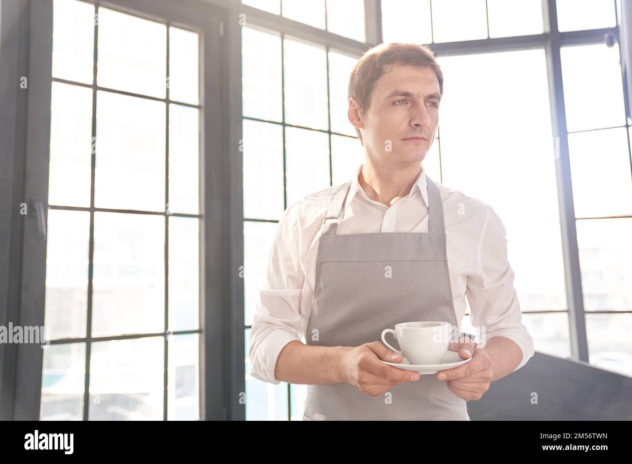 Ein männlicher Kellner in einer grauen Schürze hält eine Tasse Kaffee. Der Barista serviert eine Tasse heißen Kaffee vor dem Hintergrund eines großen Fensters mit Platz zum Kopieren. Hochwertiges Foto Stockfoto