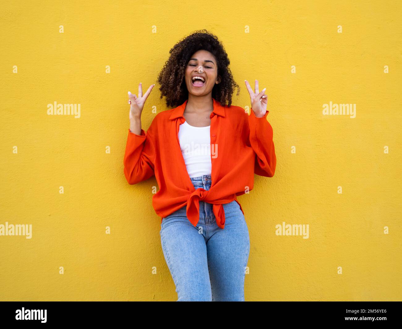 Fröhliche afroamerikanische junge Frau, die lächelt und ein Siegessymbol mit Händen über der gelben Wand macht. Eine Geste zum Siegesfeiern Stockfoto