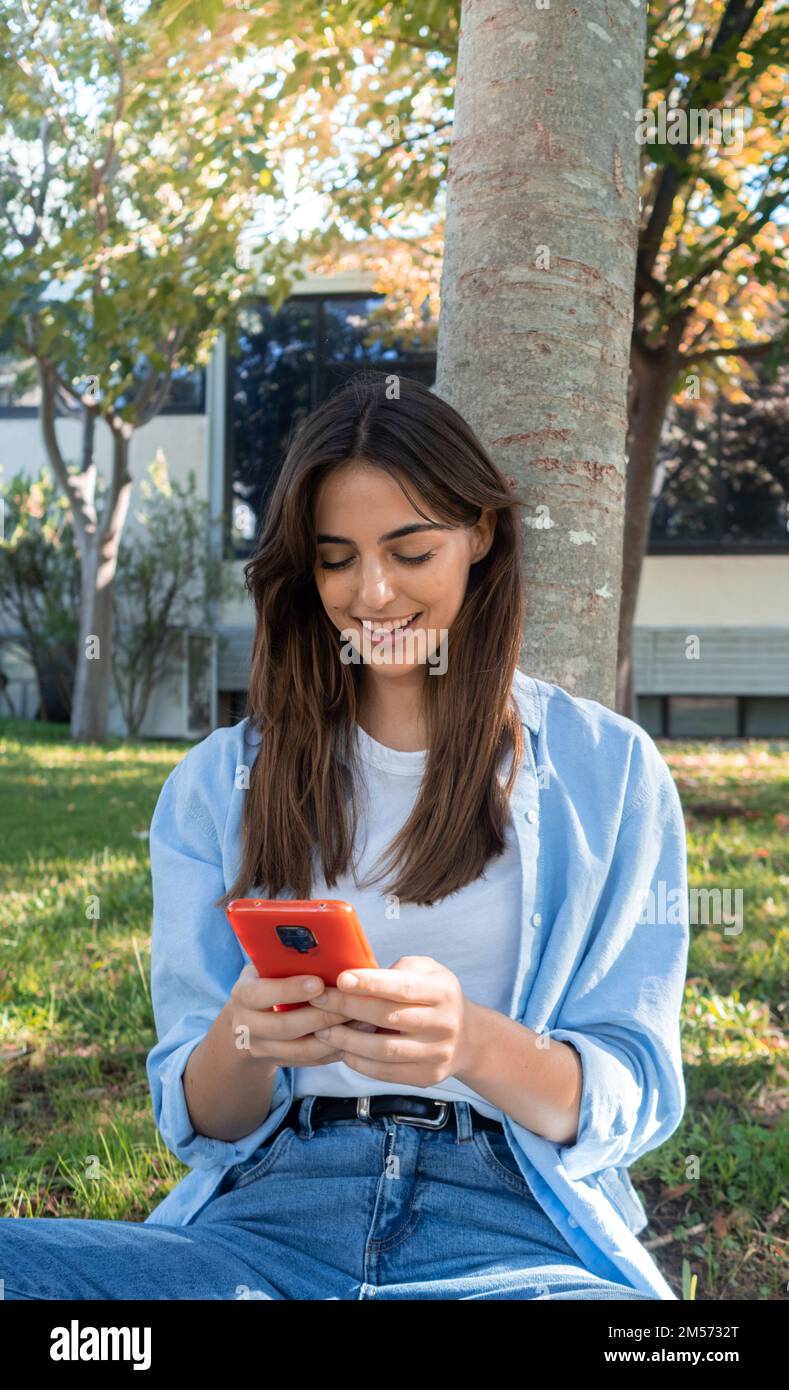 Eine lächelnde junge Frau der Millennials unterhält sich mit einem Smartphone, das in einem Park sitzt Stockfoto