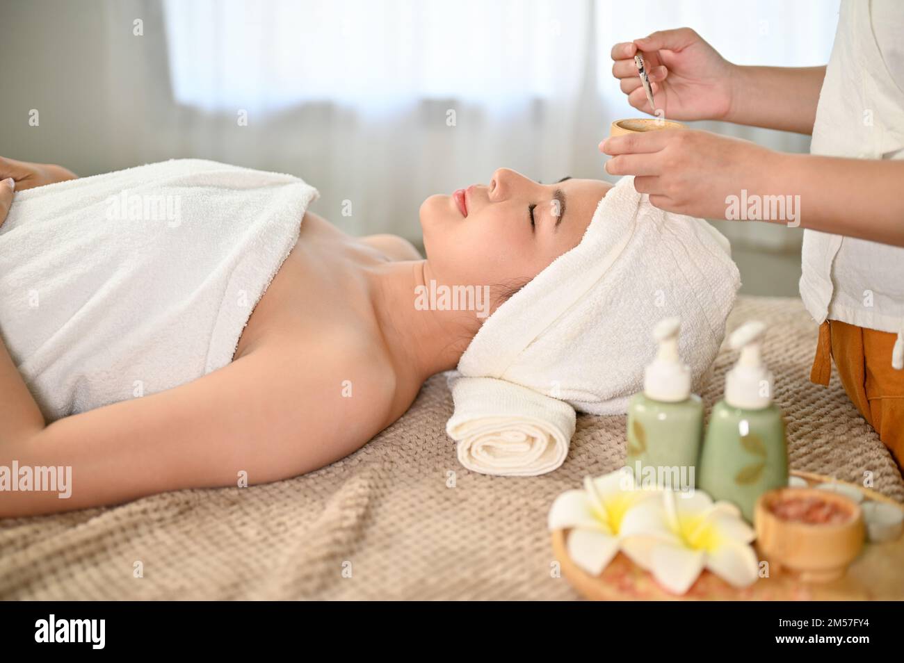 Wunderschöne asiatische Frau, die auf dem Massagetisch liegt und eine Gesichtsbehandlung mit Tonmaske von einem Spa-Mitarbeiter erhält. Seitenansicht Stockfoto