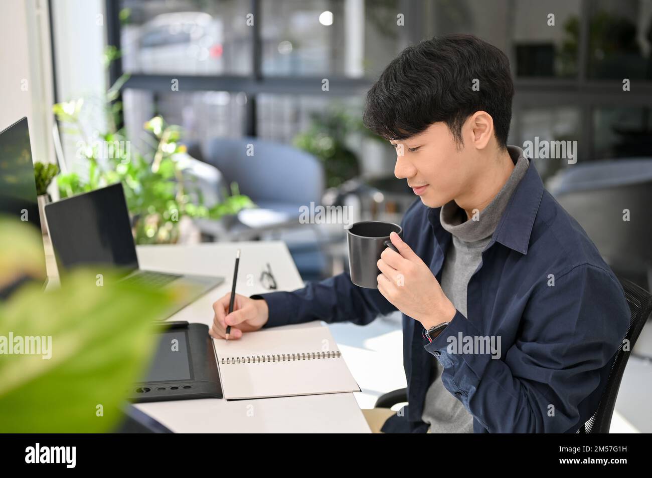 Attraktiver und kluger junger asiatischer Büroangestellter, der sich auf seine Aufgabe konzentriert, eine Liste auf seinem Notizblock erstellt, während er morgens Kaffee an seinem Schreibtisch trinkt. SI Stockfoto