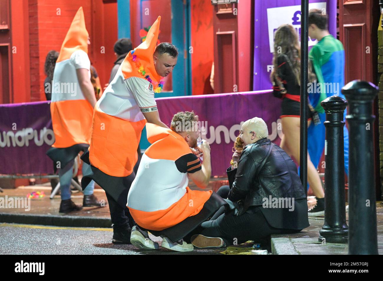 King Street, Wigan, 27. 2022. Dezember. - Reveller stolperten nach Hause und ruhten sich gegen Gebäude und auf Gehwegen aus, nach einem schönen Abend am Boxing Day, der alljährlichen Fancy Dress Night in Wigan. Ein paar Verkehrskegel stellten sicher, dass es einer Frau gut ging, als sie auf einem Bordstein saß. Ein Mann in einem Frauenkleid, Freddie Mercury-Stil, wurde angeschrien und von einer anderen Frau außerhalb von Popworld weggestoßen. Einige nahmen sich ein Essen mit, um die Nacht davor zu genießen, als sie auf Taxis warteten. Stockfoto