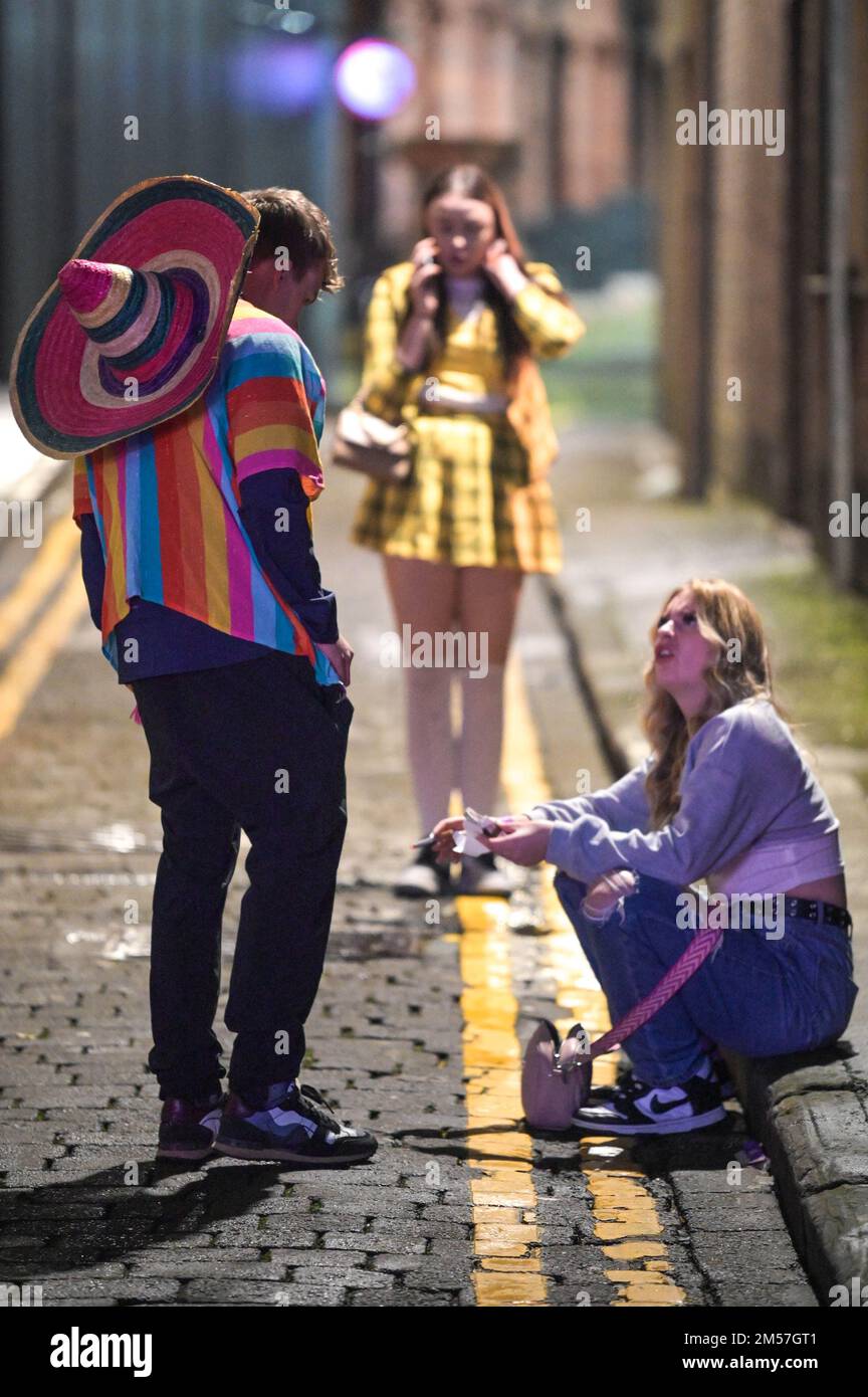 King Street, Wigan, 27. 2022. Dezember. - Reveller stolperten nach Hause und ruhten sich gegen Gebäude und auf Gehwegen aus, nach einem schönen Abend am Boxing Day, der alljährlichen Fancy Dress Night in Wigan. Ein paar Verkehrskegel stellten sicher, dass es einer Frau gut ging, als sie auf einem Bordstein saß. Ein Mann in einem Frauenkleid, Freddie Mercury-Stil, wurde angeschrien und von einer anderen Frau außerhalb von Popworld weggestoßen. Einige nahmen sich ein Essen mit, um die Nacht davor zu genießen, als sie auf Taxis warteten. Stockfoto