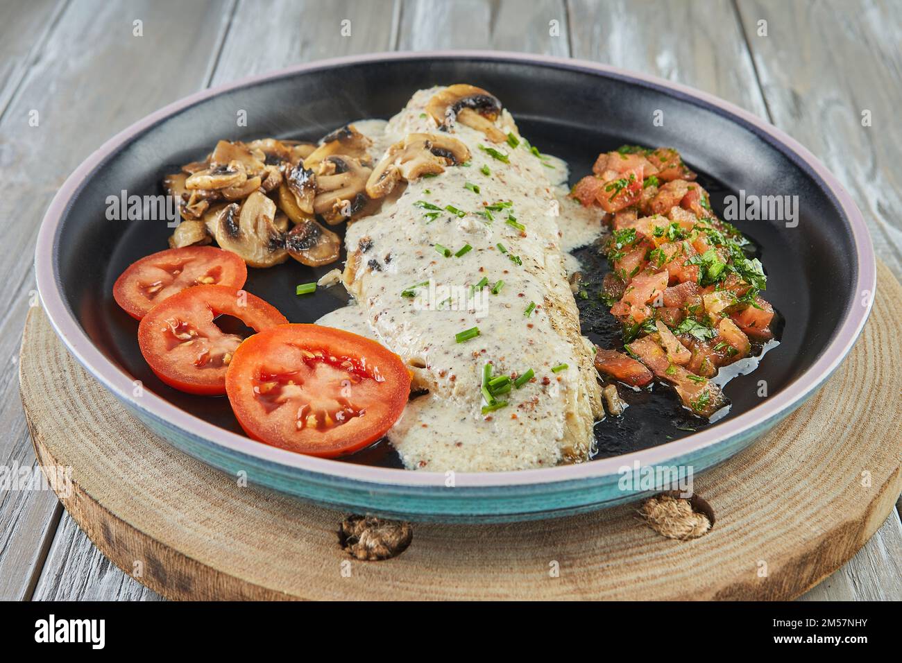 Wolfsbarsch mit Senf, Pilzen und Tomaten auf schwarzem Teller Stockfoto