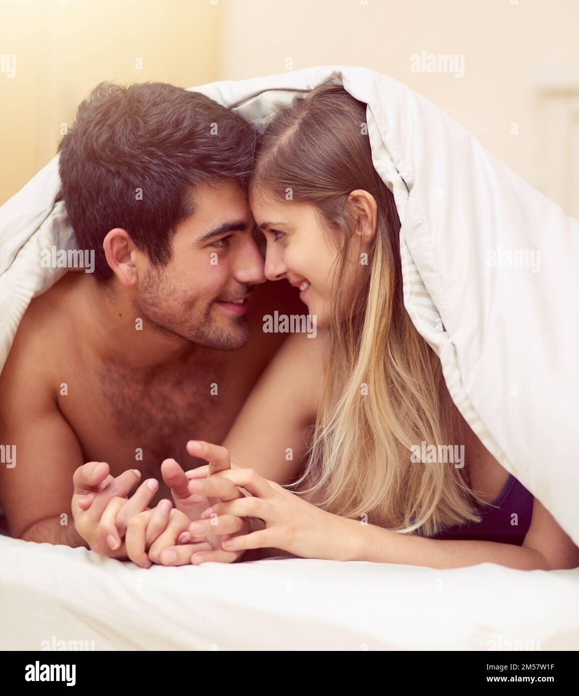 Sich in den Augen der anderen zu verirren. Ein liebevolles junges Paar, das sich anlächelt, während es unter einer Decke liegt. Stockfoto