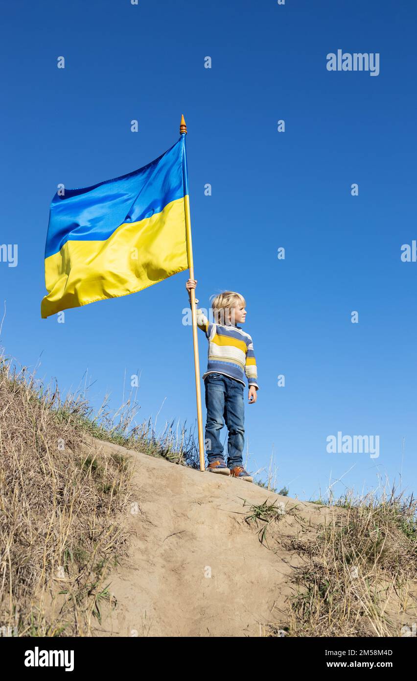 An sonnigen Tagen steht der Junge auf dem Hügel gegen den blauen Himmel und hält eine große winkende blaue und gelbe Nationalflagge. Stolz, Ukrainer zu sein. Kinder gegen Krieg. st Stockfoto