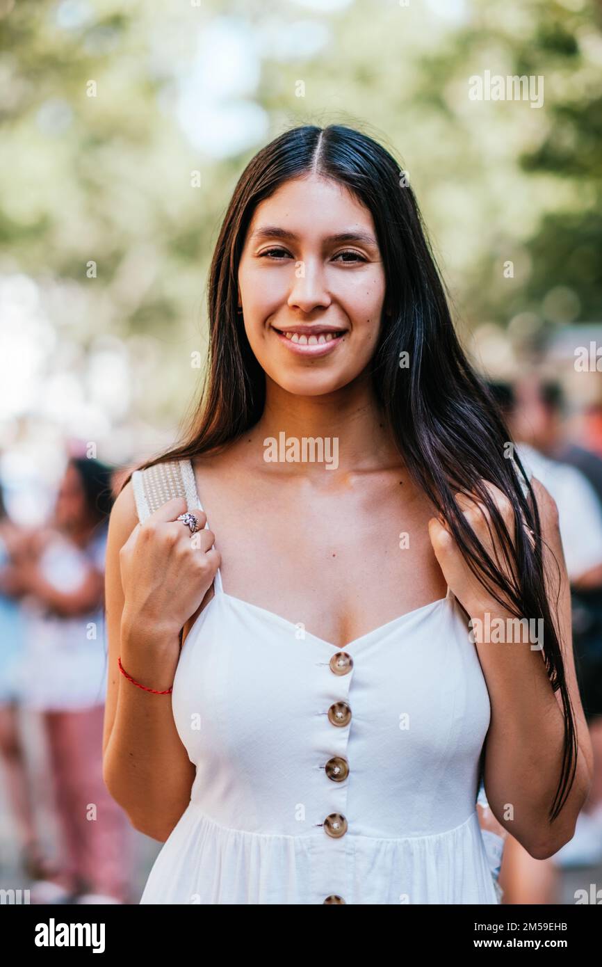 Glückliche lateinamerikanische Frau in weißem Kleid, die an einem sonnigen Sommertag in Barcelona, Spanien, Rucksackträger an den Schultern berührt und mit einem Lächeln auf den unscharfen Hintergrund des Parks in die Kamera schaut Stockfoto