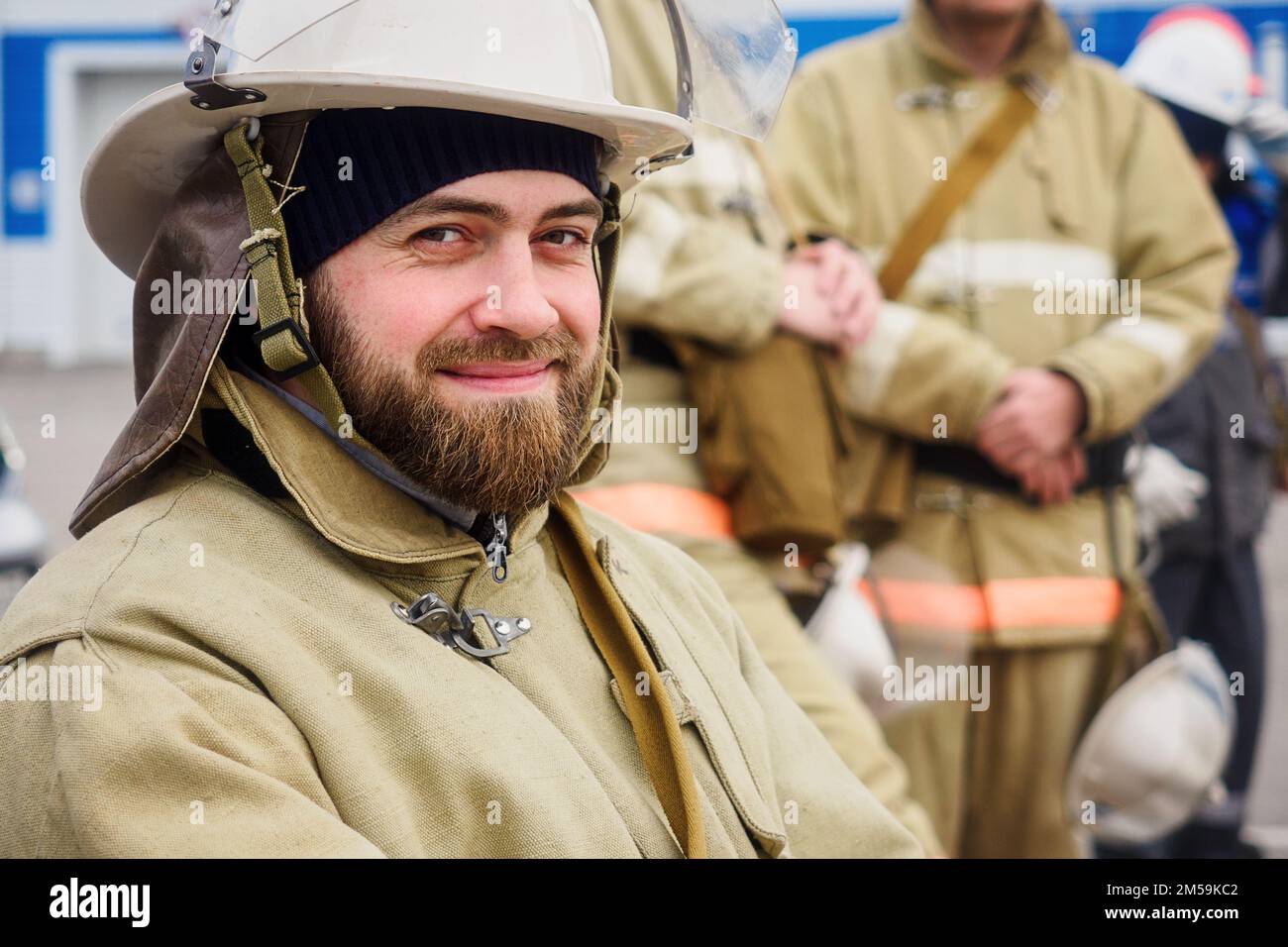 Porträt eines bärtigen Retters am Sommertag auf der Straße. Feuerwehrmann in Schutzkleidung und Helm schaut in die Kamera und lächelt. Authentischer weißer Rettungshelfer. Positive Emotionen... Stockfoto