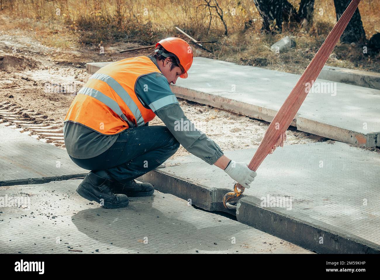 Der Schleuder legt am Sommertag Betonplatten auf die Baustelle. Arbeiter in Schutzweste und Bauhelm beaufsichtigt das Verlegen der Basis auf der Baustelle. Stockfoto