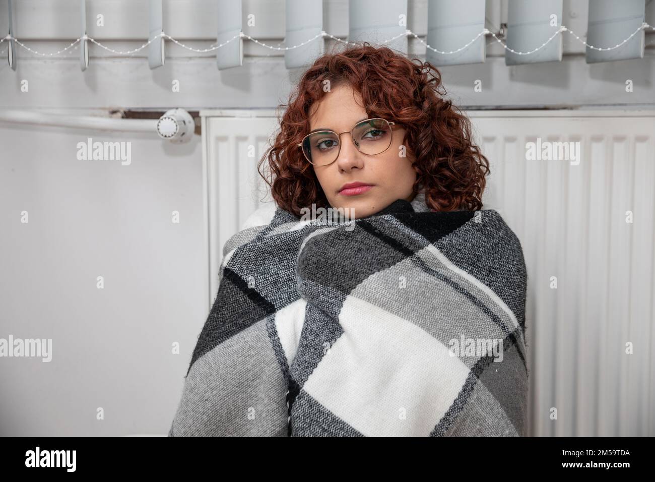 Ein Mädchen, eingewickelt in einen dicken Schal, sitzt neben einem warmen Heizkörper. Zentralheizung. Stockfoto