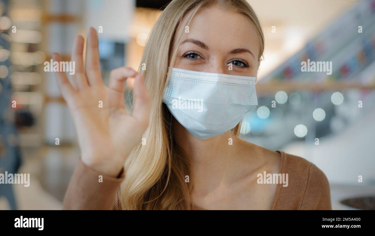 Portrait des jungen Mädchens in schützender medizinischer Maske weiße Frau zeigt Zeichen okay freudig blond schaut auf die Kamera und genehmigt Symbol Geste Stockfoto