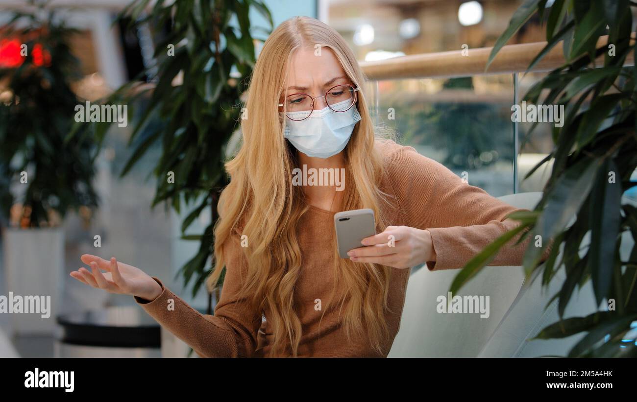 Verärgerte blonde kaukasische Frau in medizinischer Maske auf Smartphone Bildschirm unzufrieden mit schlechte Nachrichten Spam Betrug negative sms App Problem Stockfoto