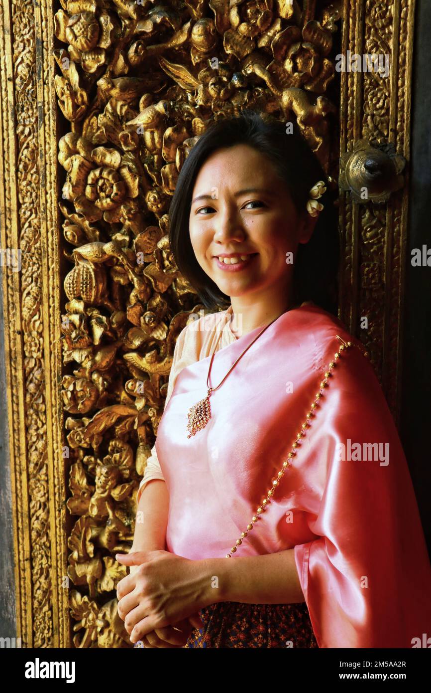 Die thailändische Dame im thailändischen Kostüm lächelt und steht an der thailändischen Holzschnitzertür Stockfoto