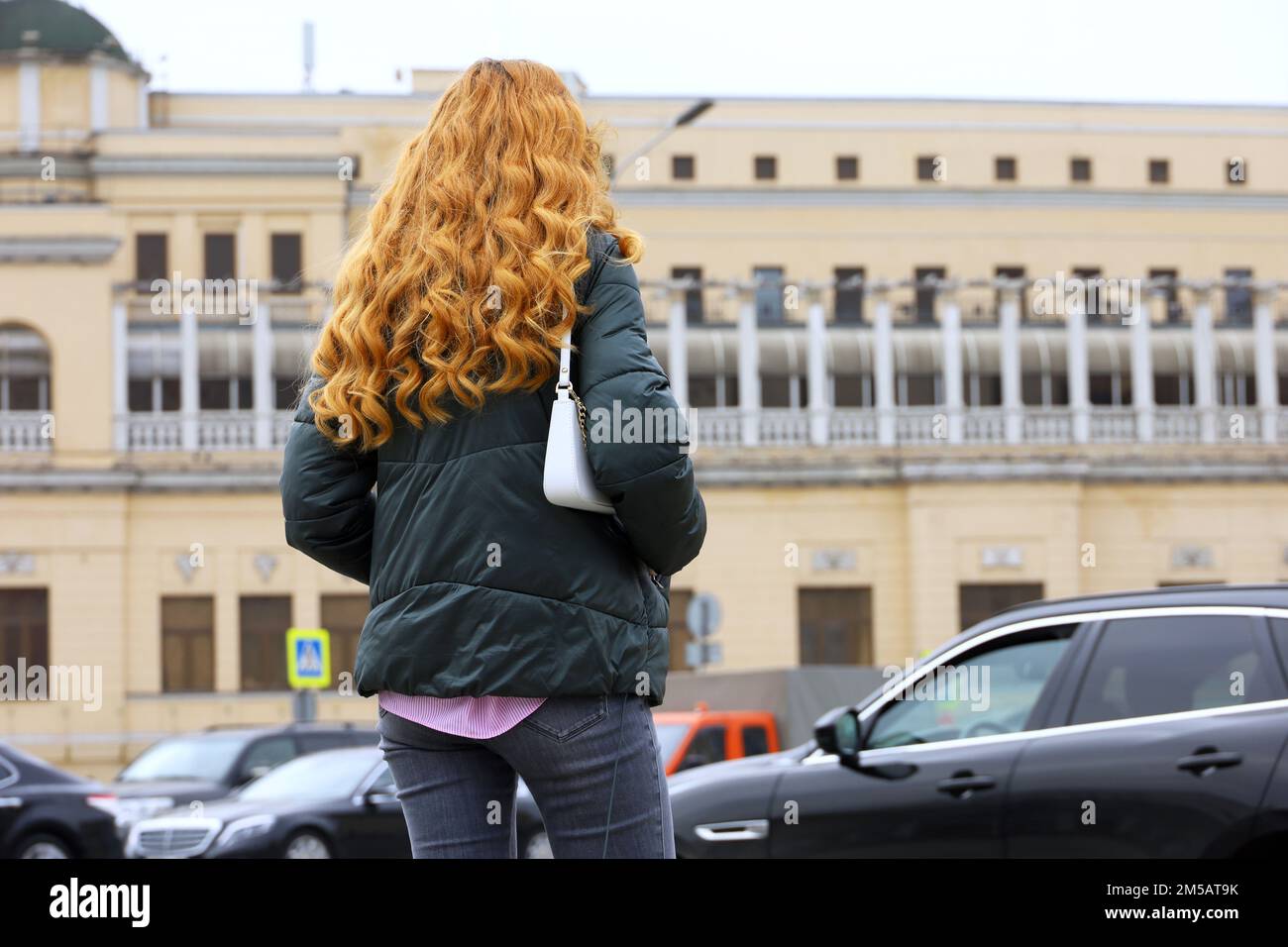 Rothaarige Frau mit langen lockigen Haaren, die Jacke und Jeans trägt und auf der Straße steht, vor dem Hintergrund eines Autos Stockfoto