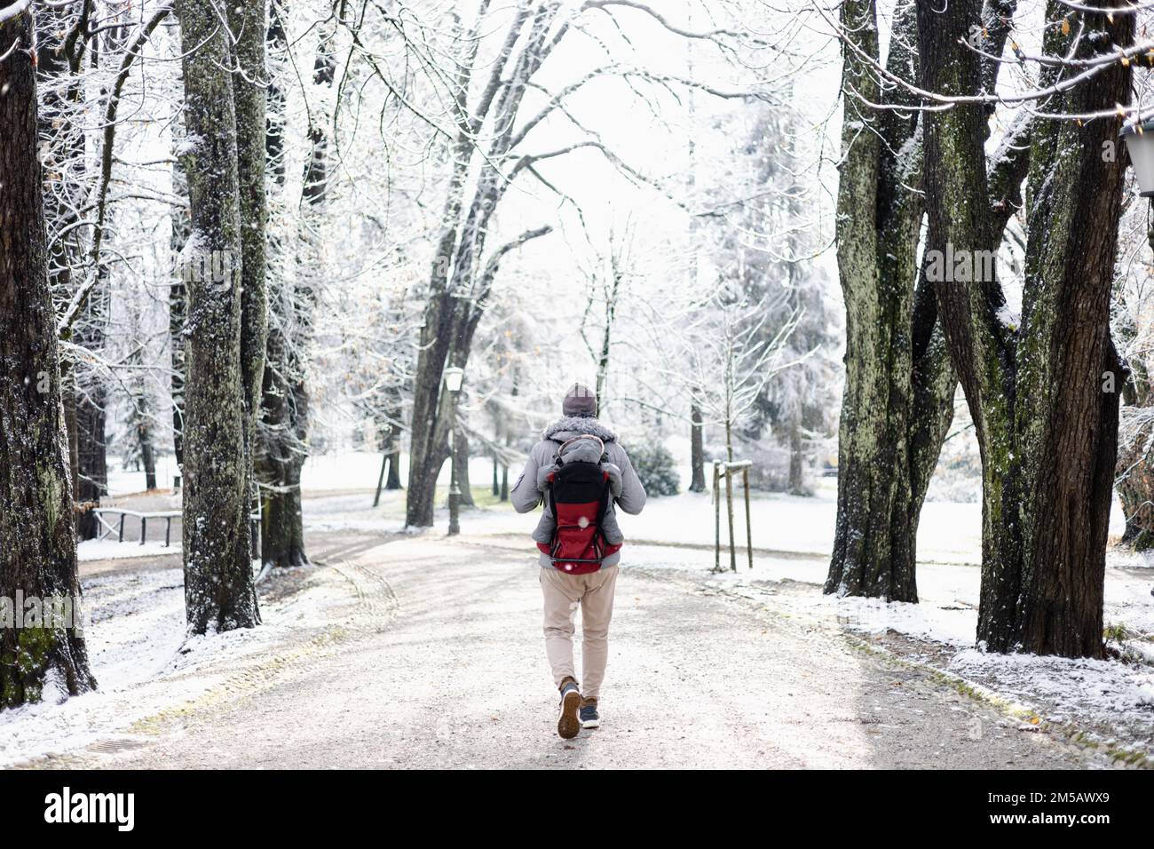 Rückansicht eines sportlichen Vaters, der seinen kleinen Sohn mit Winteranzug und Mütze im Rucksackträger im Winter im Stadtpark herumlaufen lässt. Stockfoto