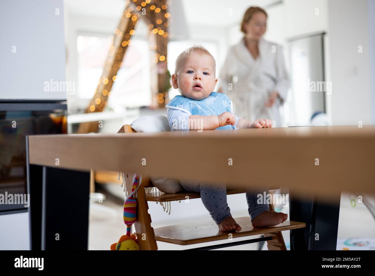 Glückliches Kind, das am Esstisch sitzt und mit seinem Spielzeug in einem traditionellen skandinavischen Designer-Hochstuhl aus Holz in einem modernen, hellen Haus spielt, das von seiner Mutter überholt wird. Stockfoto