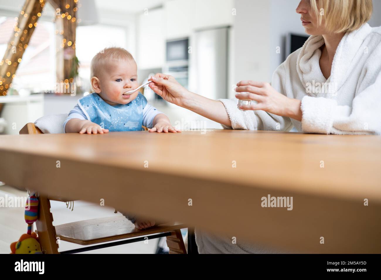 Mutter trägt einen gemütlichen Bademantel Löffel und füttert ihr Kind im Baby-Hochstuhl mit Fruchtpüree am Esstisch zu Hause. Einführungskonzept für feste Babynahrung. Stockfoto