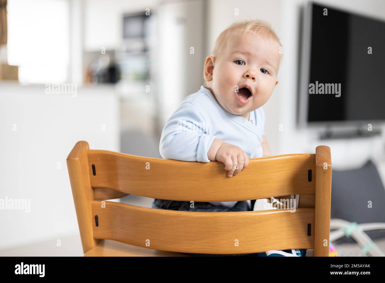 Glückliches Baby, das in einem traditionellen skandinavischen Designer-Hochstuhl aus Holz in einem modernen, hellen Zuhause sitzt. Süßes Baby Stockfoto