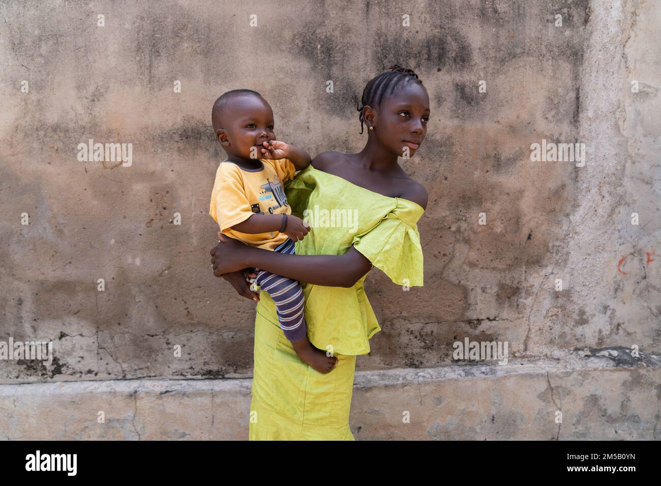 Hübsches, junges afrikanisches Mädchen in einem leuchtend gelben Kleid mit ihrem süßen kleinen Bruder in den Armen; Symbol für Familienbande Stockfoto