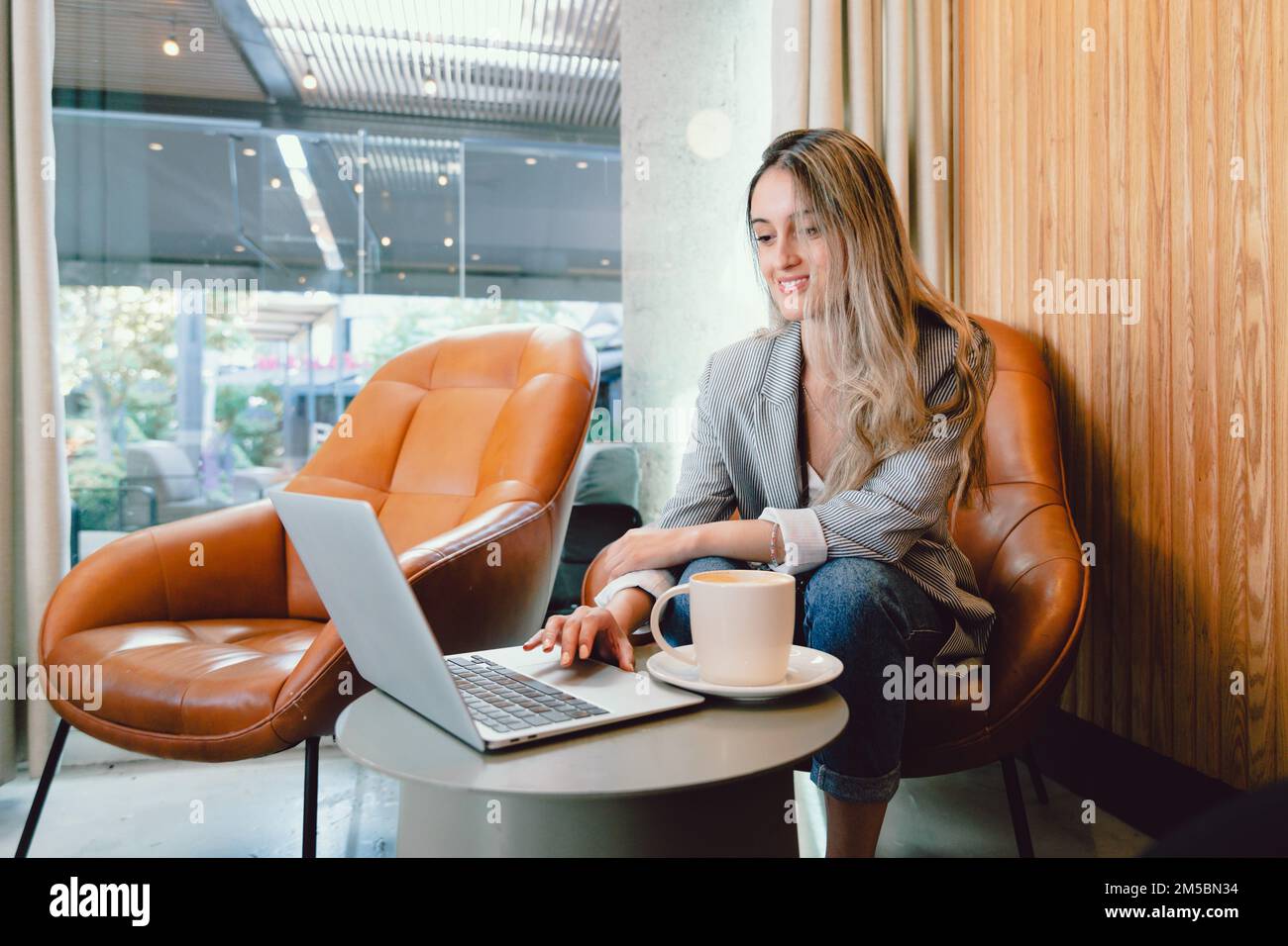 Friedliche Geschäftsfrau der Jahrtausendwende, die einen Computer benutzt, mit einer Tasse Kaffee am modernen Arbeitsplatz. Stockfoto