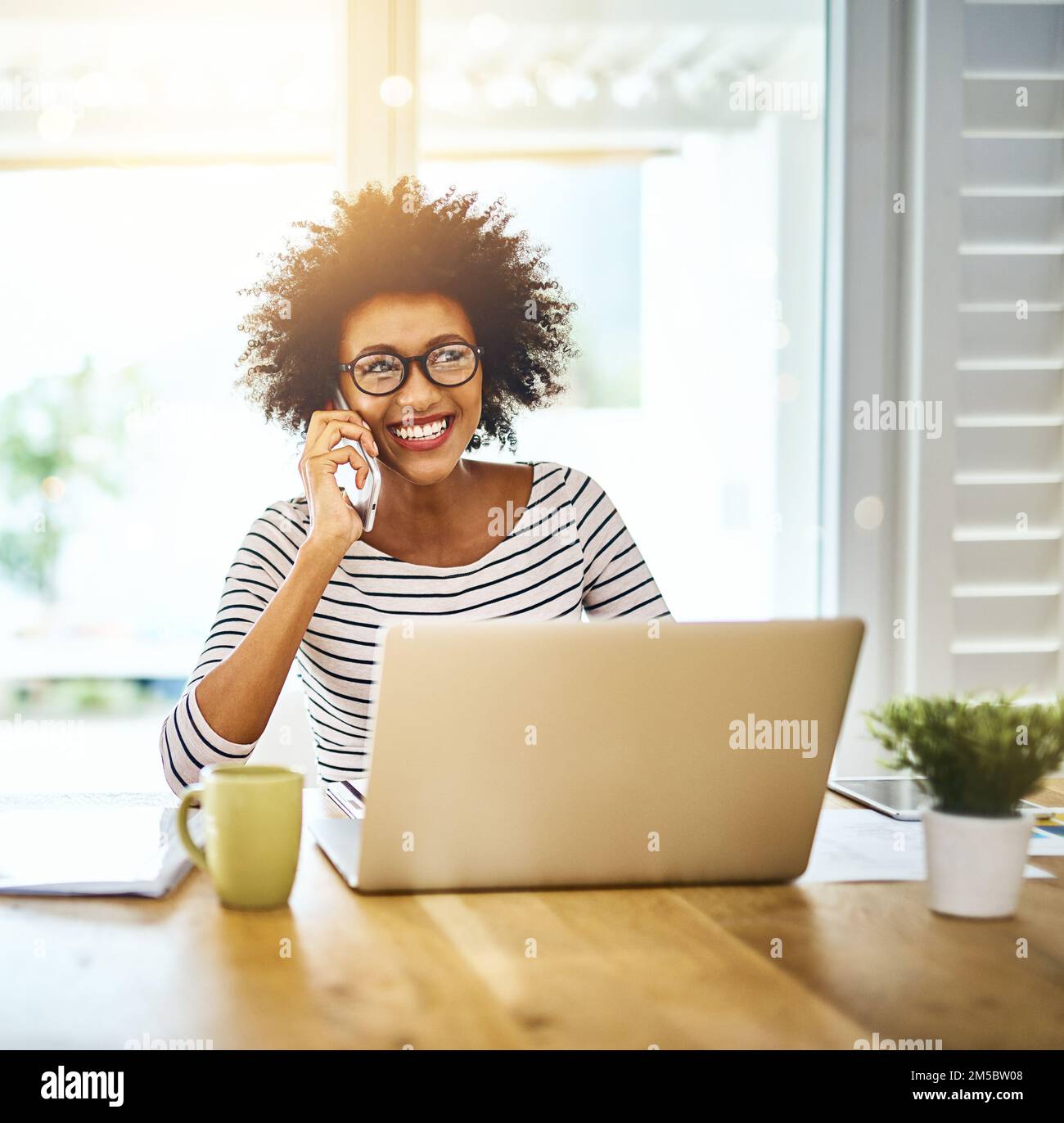 Geschäftliche Anrufe von zu Hause aus tätigen. Porträt einer fröhlichen jungen Frau, die mit ihrem Telefon spricht, während sie zu Hause an einem Notebook arbeitet. Stockfoto