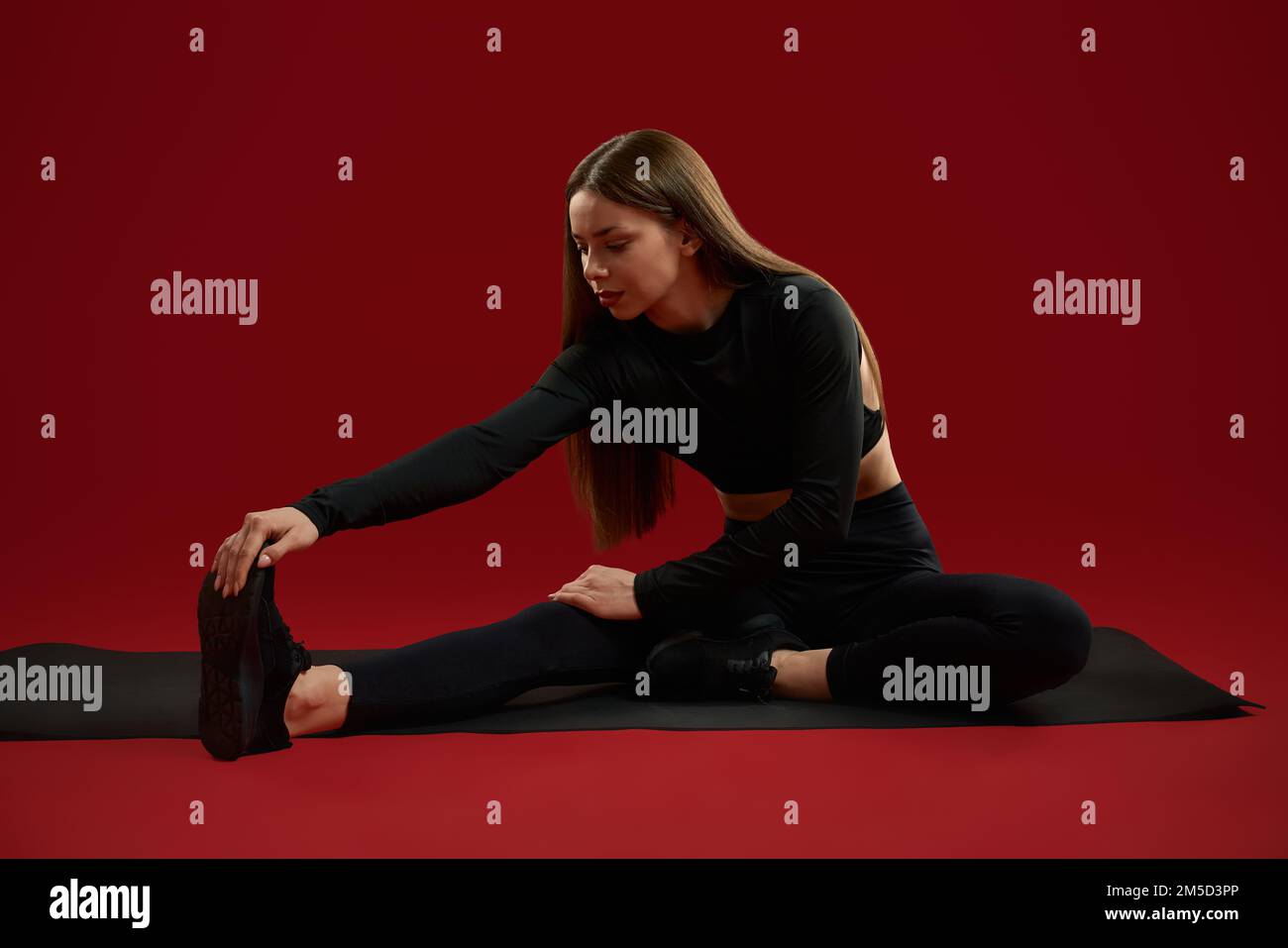 Flexibilität brünette Frau in Schwarz für Stretchübungen an den Beinen und Füße an der Socke ziehen. Ziemlich ernste weibliche Haltung mit Bein in der Seite, Training im roten Studio. Konzept des sportlichen Lebensstils. Stockfoto