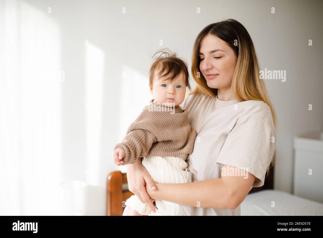 Süßes kleines Mädchen unter 1 Jahren an den Mutterhänden tragen Sie einen lässigen Strickpullover und eine Hose im Schlafzimmer. Schau in die Kamera. Mutterschaft. Stockfoto