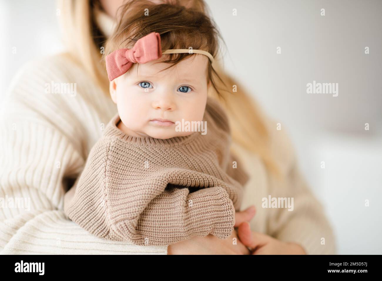 Süßes kleines Mädchen unter 1 Jahren an den Händen der Mutter trägt im Schlafzimmer einen Strickpullover und eine Hose. Schau in die Kamera. Mutterschaft. Stockfoto