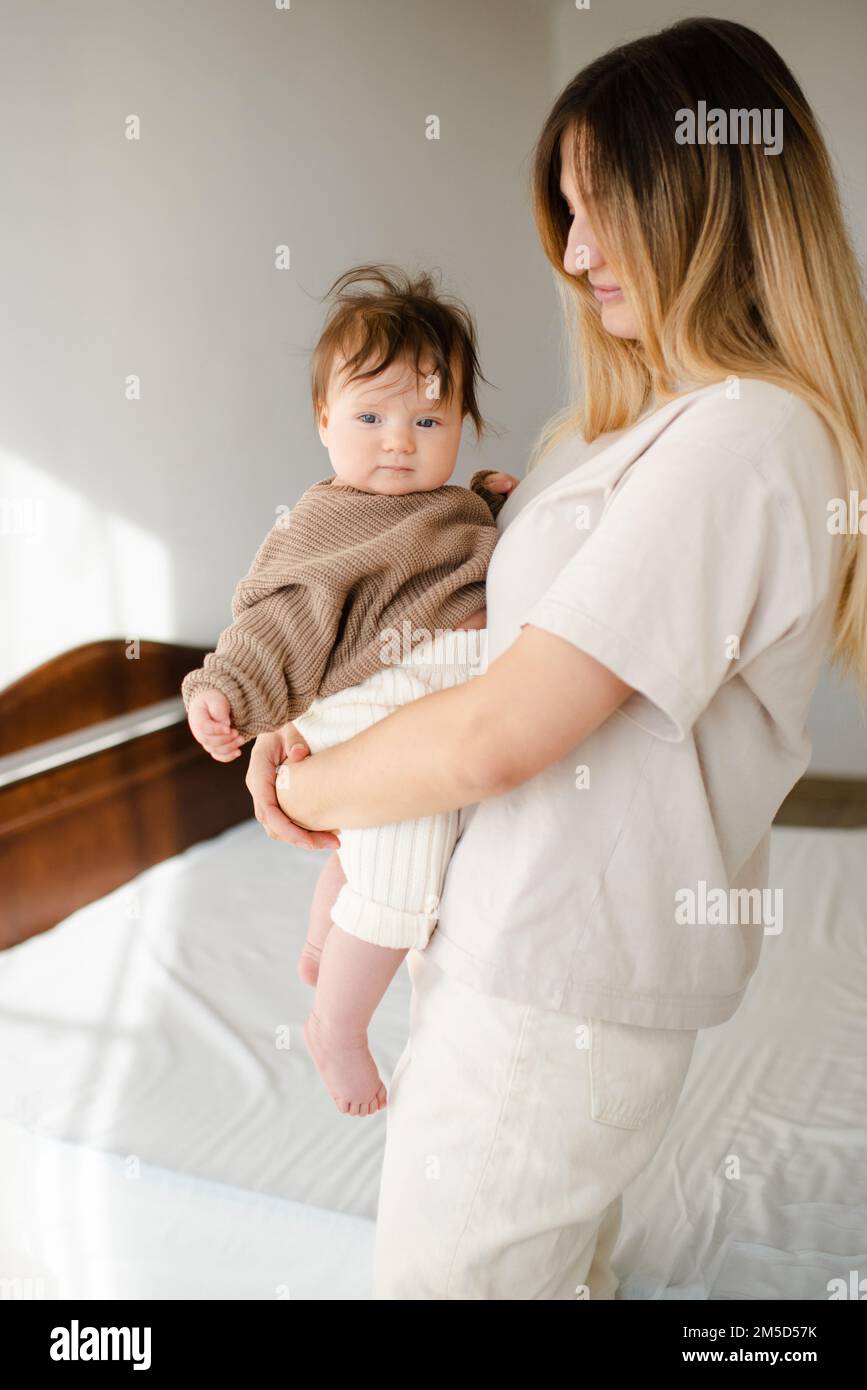 Süßes kleines Mädchen unter 1 Jahren an den Mutterhänden tragen Sie einen lässigen Strickpullover und eine Hose im Schlafzimmer. Schau in die Kamera. Mutterschaft. Stockfoto