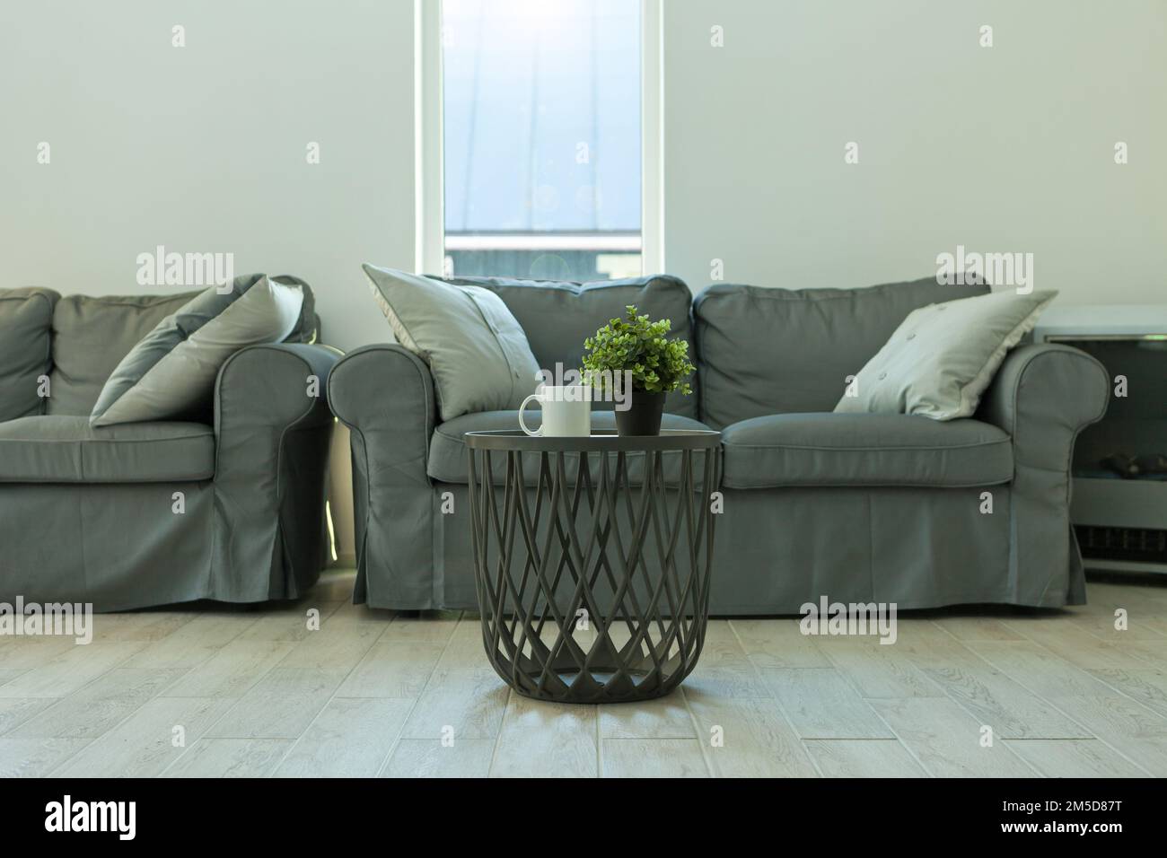 Graue Innenausstattung mit Fenster. Schrank mit Anlage. Graues Sofa im einfachen Wohnzimmer. Tisch mit Becher Stockfoto