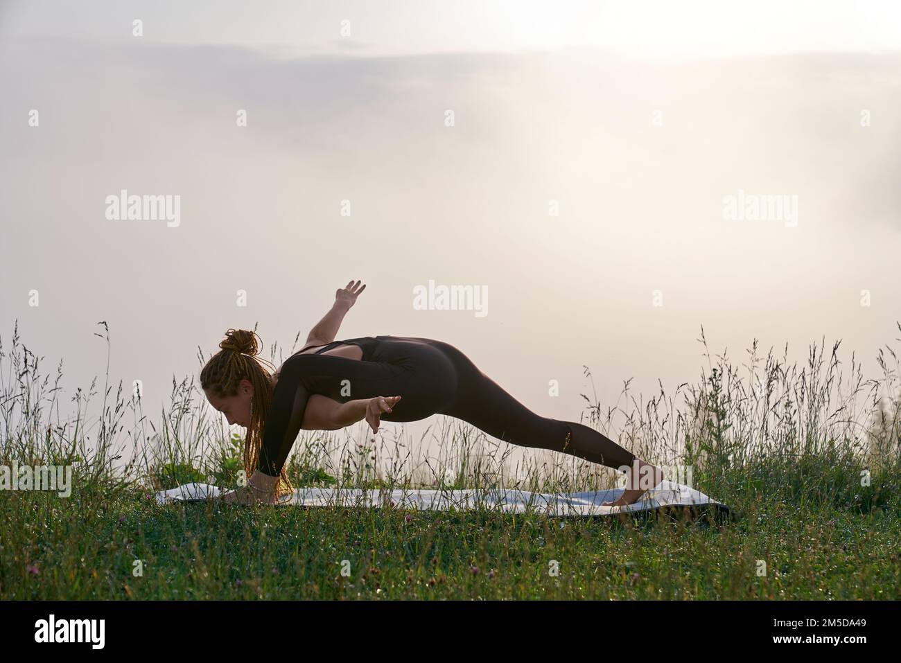 Aktive Frau mit Zöpfen, die im Freien Yoga-Übungen auf der Matte macht. Gesunde und fit Frau in Sportkleidung, die Freizeit für das Training an der frischen Luft verbringt. Stockfoto