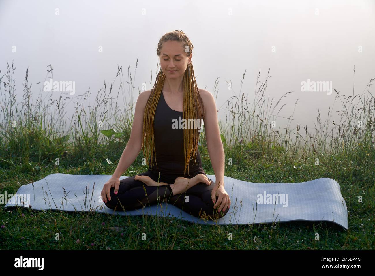 Friedliche junge Frau mit geflochtenem Haar, die in Lotus meditiert, posieren in grüner Natur. Gesunde Frau, die morgens im Freien Harmonie genießt. Stockfoto