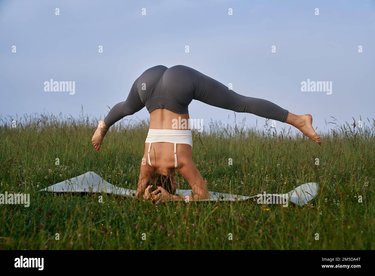 Sportliche junge Frau in Sportkleidung, die sich die Beine streckt, während sie im Freien Kopfständerübungen macht. Regelmäßiges Workout für einen gesunden und gesunden Körper. Stockfoto