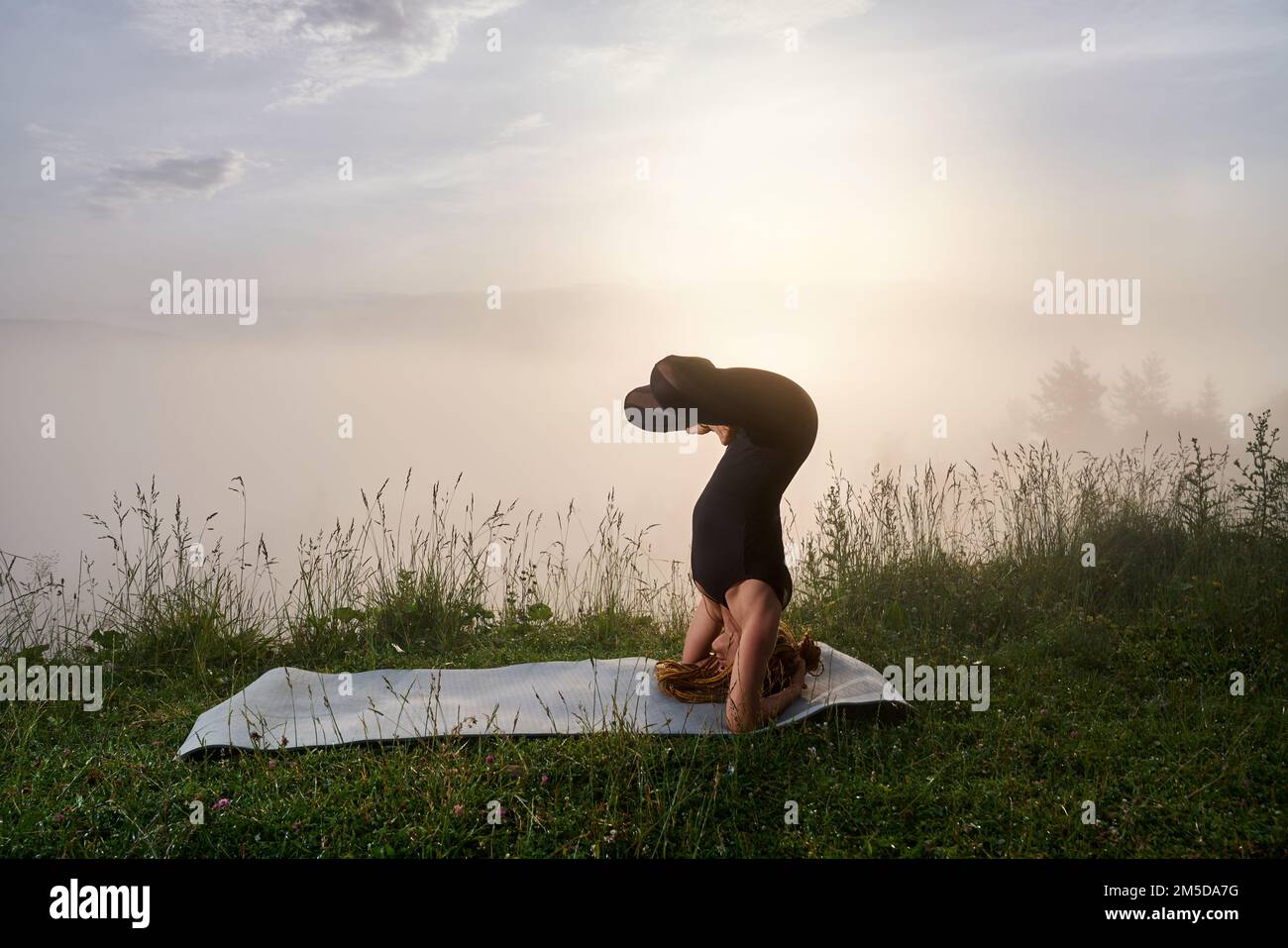 Eine starke junge Frau in schwarzer Sportkleidung, die im Freien mit dem Kopfständer trainiert. Sommersonnenaufgang über den Bergen im Hintergrund. Stockfoto