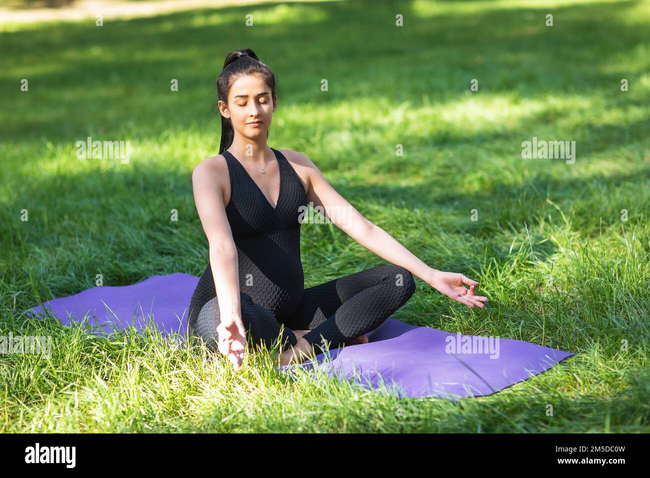 Eine junge schwangere Frau meditiert in Lotusposition mit Jnana mudra, übt Yoga auf einer Matte im Park in einem schwarzen, einteiligen Jumpsui Stockfoto