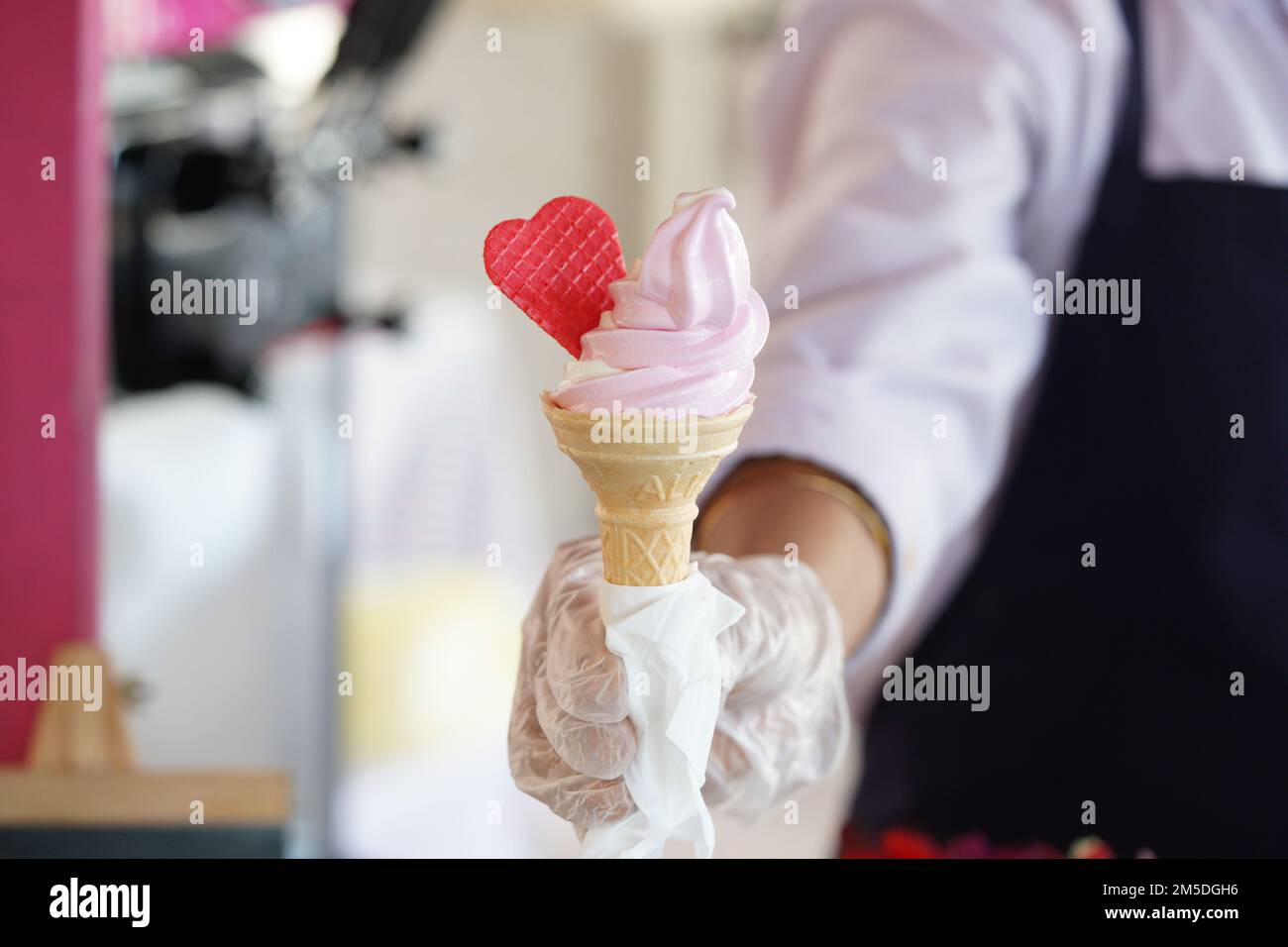 Ein Arbeiter mit Handschuhen, der eine Eiskugel mit einem Mix aus Rosa und Vanille hält. Rosa Eiscreme mit rotem Herzen. Stockfoto