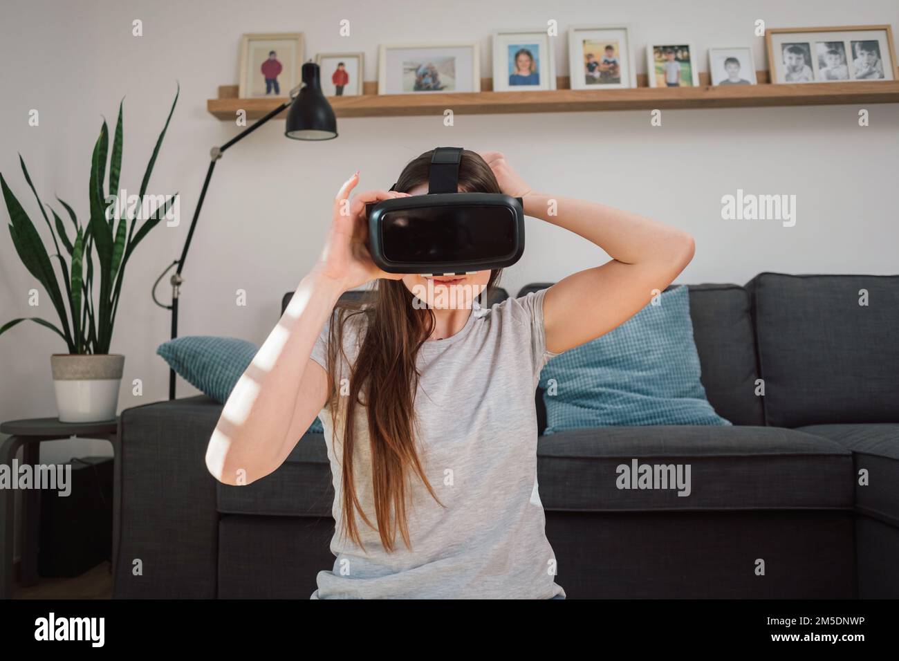 Eine junge Frau trägt eine Virtual-Reality-Brille, erkundet die erweiterte Welt, interagiert mit der digitalen Schnittstelle, während sie im Wohnzimmer sitzt Stockfoto