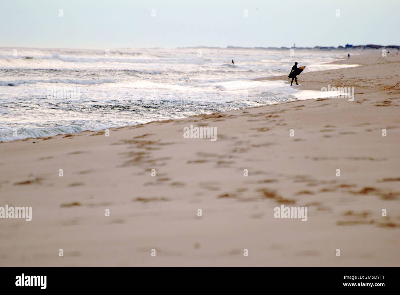 Ein einsamer Surfer trägt sein Surfbrett von den Wellen des Ozeans über einen verlassenen Strand in der Nebensaison des Winters Stockfoto
