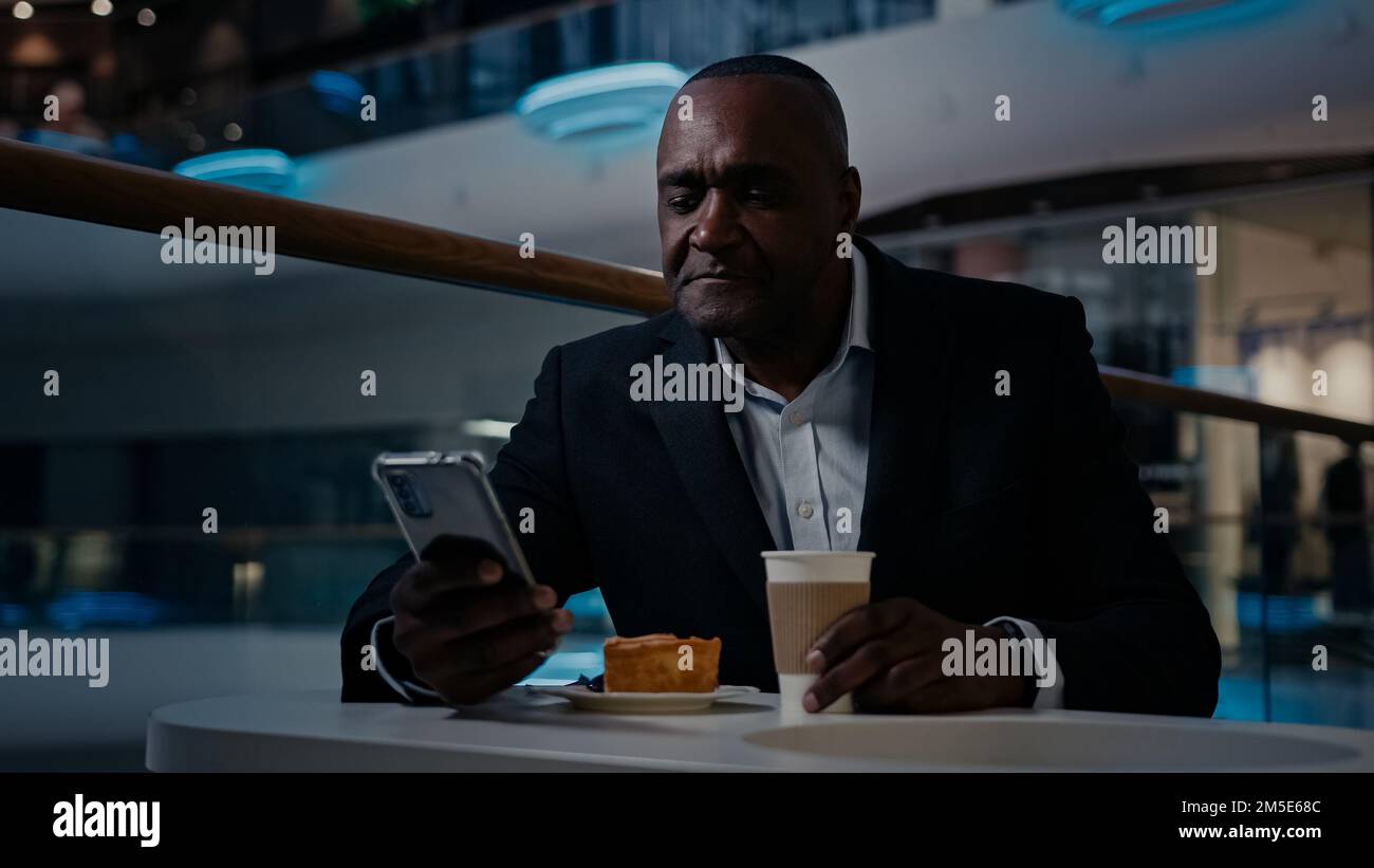 Afrikanischer erwachsener Geschäftsmann mittleren Alters, 50s ethnischer Mann im Anzug Arbeitgeber Unternehmer sitzt am Tisch im Café trinken Kaffee trinken Tee surfen Stockfoto