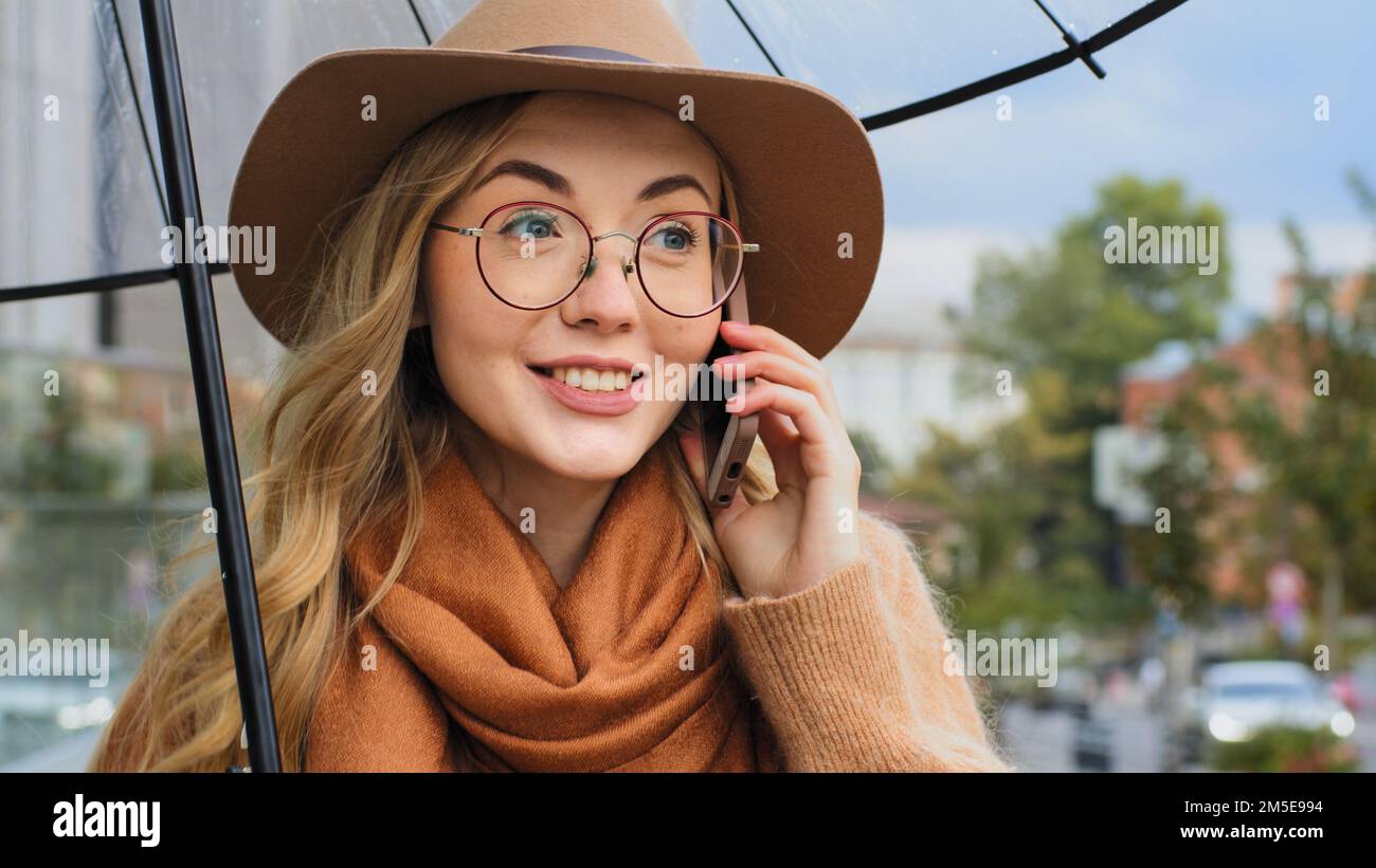 Attraktive junge Frau mit Schirm beantwortet Anrufe auf der Straße im Regen, kluges Mädchen kommuniziert enthusiastisch, emotionale weiße Dame Stockfoto