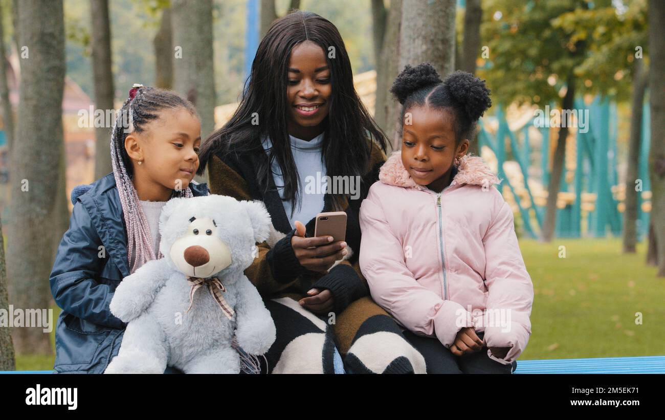 Freundliche Familie, die auf einer Bank im Park sitzt, junge mutter, die auf ihr Handy schaut, Töchter, Kind, das einen Teddybär hält, afroamerikanische Mutter kommuniziert mit ihr Stockfoto