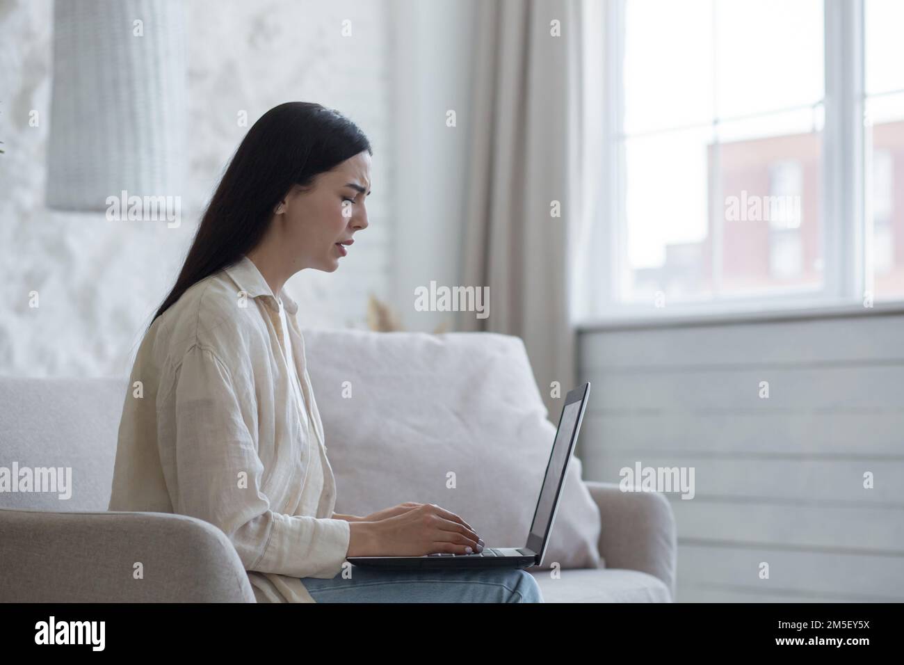 Junge verärgerte Frau erhielt schlechte Nachrichten, Dokumente, Scheidungsschreiben per Online-Mail. Sie sitzt zu Hause traurig auf dem Sofa, hält einen Laptop und weint. Stockfoto