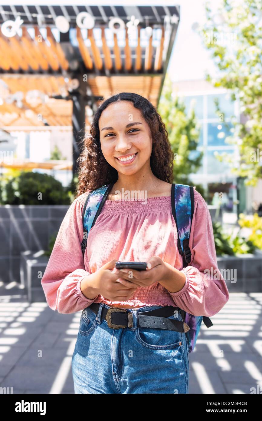 Lateinische Studentin, die lächelt und ihr Handy benutzt, um außerhalb des Campus in der Freizeit zu sitzen Stockfoto