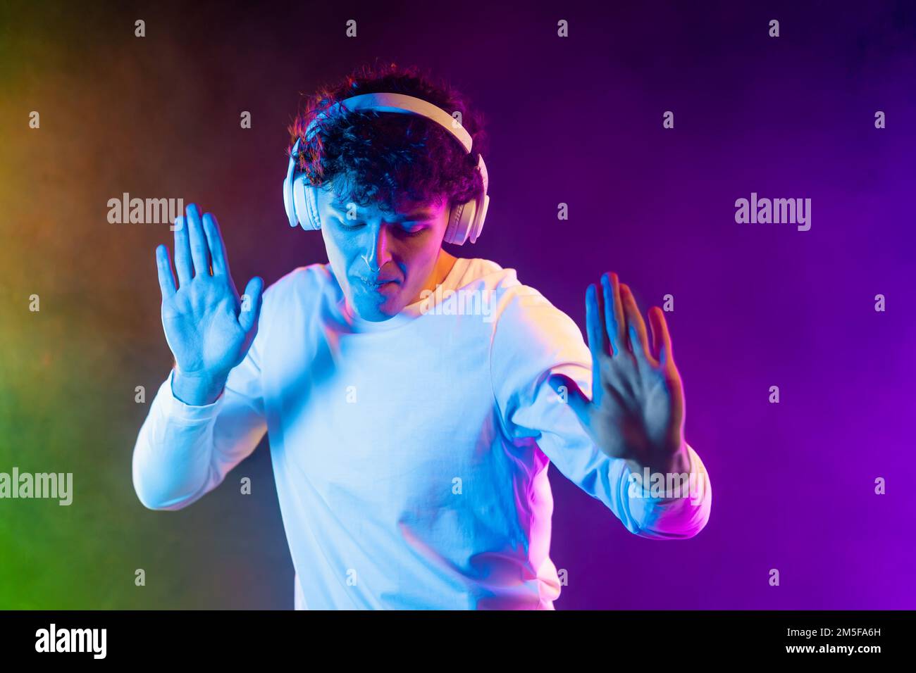 Positiver Mann, der Musik mit Kopfhörern hört, auf dunklem Neonhintergrund tanzt. Stylischer Schüler, der das Leben genießt, aktive Energie, inspirierter Tanz Stockfoto