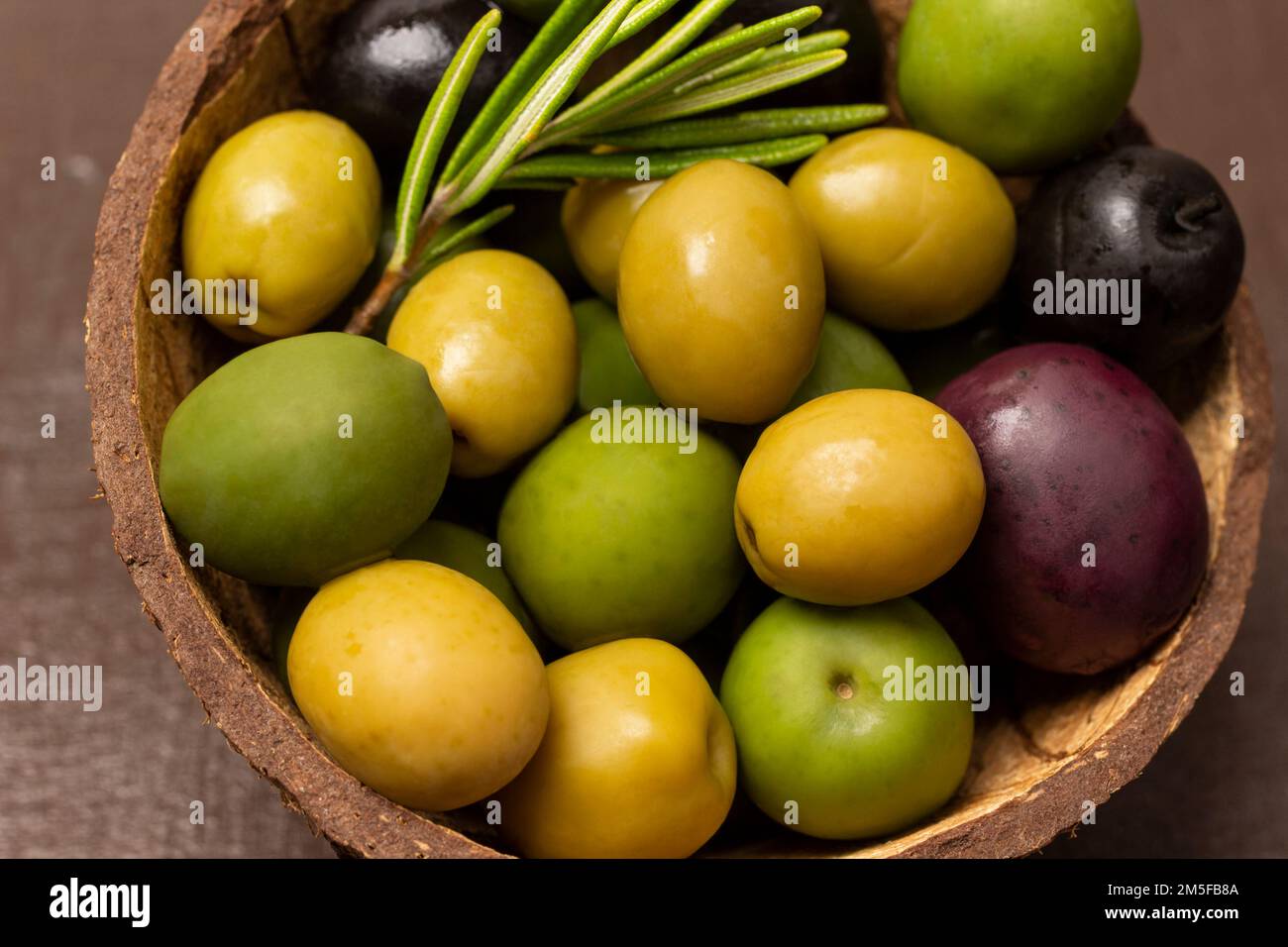 Mehrfarbige Oliven und ein Zweig Rosmarin in einer Kokosnussschale. Schließen. Stockfoto