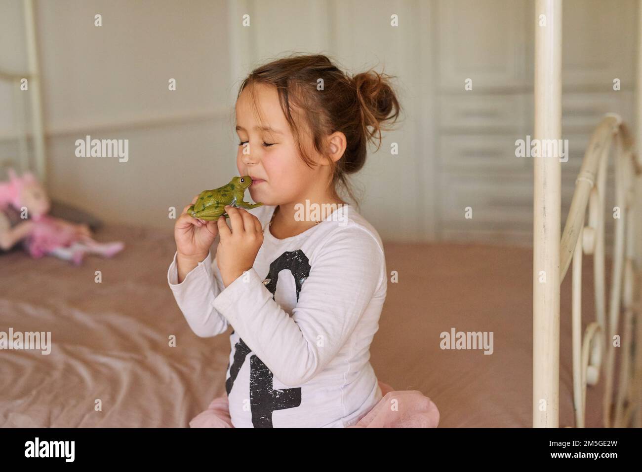 Vielleicht verwandelt es sich in einen kleinen Prinzen. Ein kleines Mädchen, das zu Hause einen Spielzeugfrosch küsst. Stockfoto