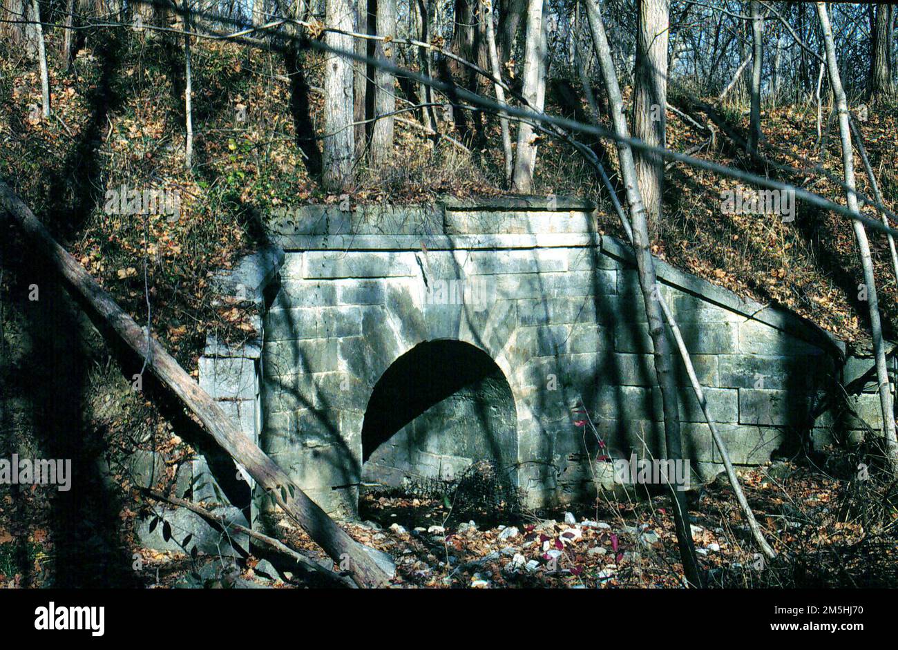 Historische Nationalstraße - Ein Steinbogen. Hohe Bäume haben ihre Herbstblätter über einen verlassenen Abschnitt der Old National Road fallen lassen. Ein Trapezbogen markiert, wo einst Reisende vorbeigingen. Östlich von Marshall, Illinois Stockfoto
