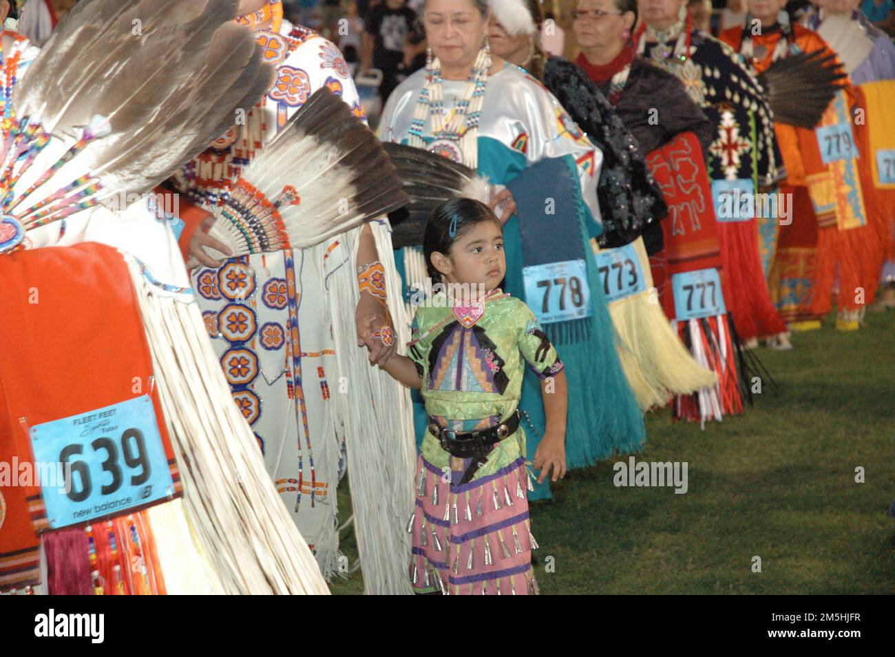 Cherokee Hills Byway - Inter Tribal Pow Wow. Ein kleines Indianermädchen, gekleidet in voller Pow-Wow-Kleidung, hält die Hand einer der Ältesten, während viele Ältere stehen, gekleidet in vollen Pow-Wow-Regalien, wie bunten Kleidern mit aufwendiger Perlenverzierung. Sie sorgen sich um wunderschöne Schals und Fans aus Federn. Jedes ist mit handgefertigtem indianischem Schmuck geschmückt. Lage: Oklahoma Stockfoto