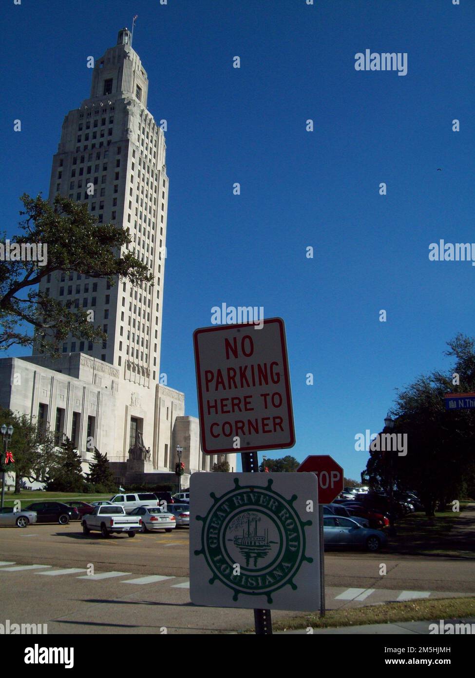 Great River Road - Schild auf der Great River Road am Louisiana State Capitol. Der Louisiana State Capitol Complex mit seinem 34-stöckigen Art Deco Statehouse steht stoisch vor einem blauen Himmel, während ein Schild mit dem Great River Road Pilot Wheel Logo im Schatten vor dem Gebäude steht. Baton Rouge, Louisiana (30,458° N 91,179° W) Stockfoto