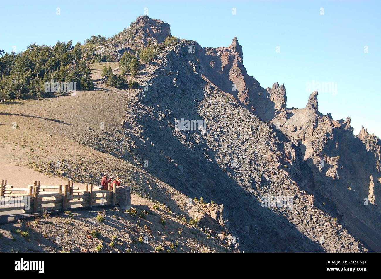 Volcanic Legacy Scenic Byway - Devil's Backbone vom Watchman Overlook. Die Leute am Watchman Overlook stehen hinter einem Geländer und schauen auf den See darunter. Über ihnen rammen die zerklüfteten Lavazähne der Devil's Backbone Formation am Rand des Crater Lake in den Himmel. Lage: Crater Lake National Park, Oregon (42,946° N 122,169° W) Stockfoto